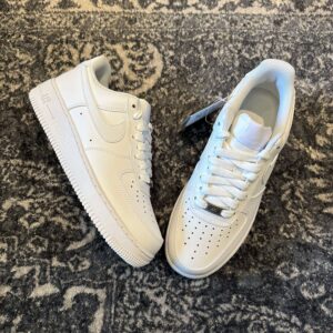 Pair of Nike Air Force 1 Low White sneakers on a textured grey rug; iconic white synthetic leather upper with perforated toe box, side view of the shoe highlighting the clean white midsole, and front view showing the laces and metallic 'AF1' lace dubrae.