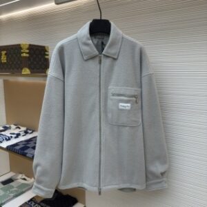 A front-view studio shot of a CD-logo Light Grey Fleece Overshirt hanging on a black hanger against a white textured wall. The overshirt features a full-length silver zipper, a classic collar, and a unique zippered chest pocket adorned with a white "Christian Dior" brand patch. In the background, a luxury boutique setting is visible, including a Louis Vuitton monogram trunk and folded designer knitwear on glass shelving.
