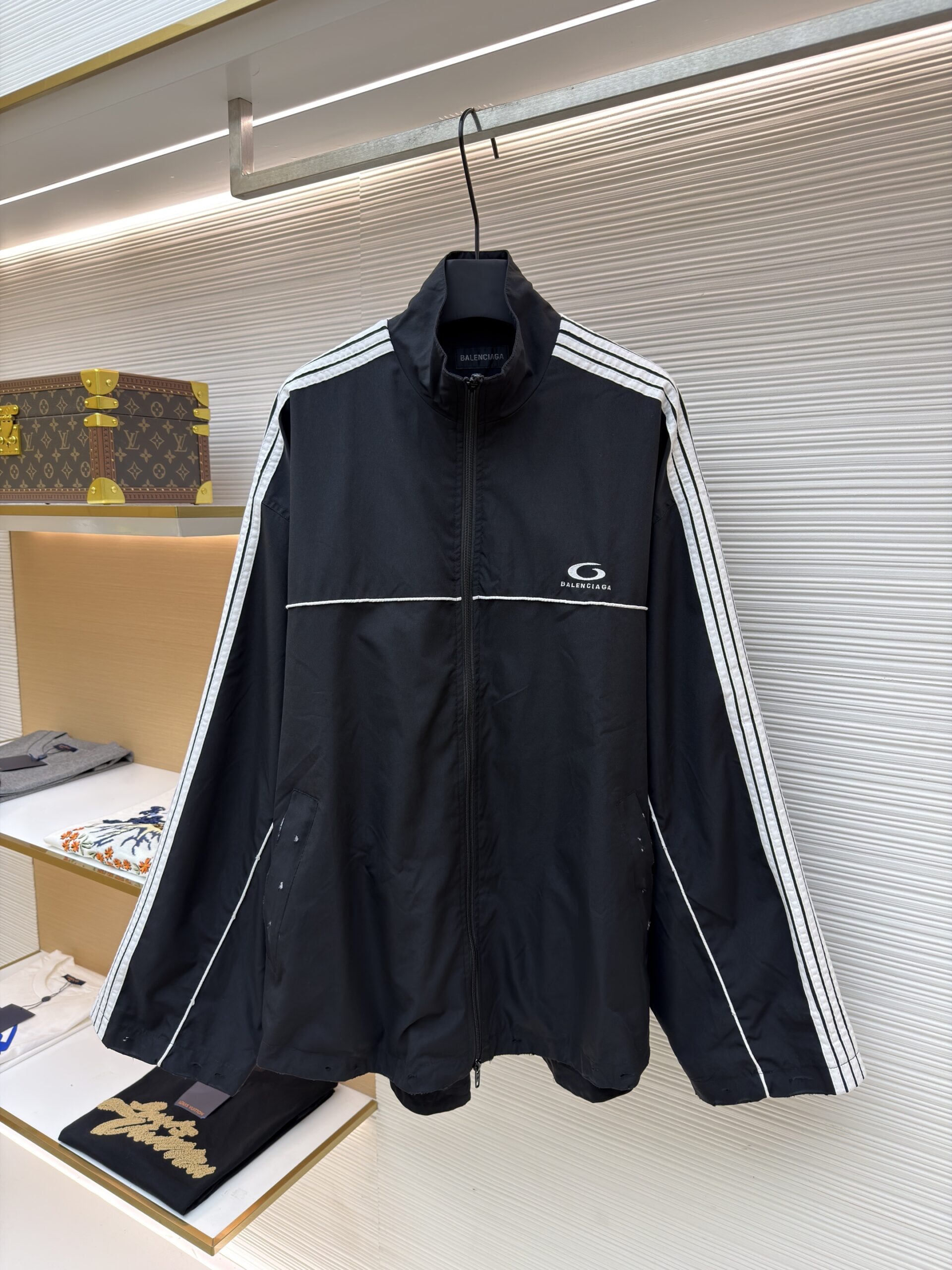 A black Balenciaga oversized track jacket with white stripes and a back logo hanging in a boutique.
