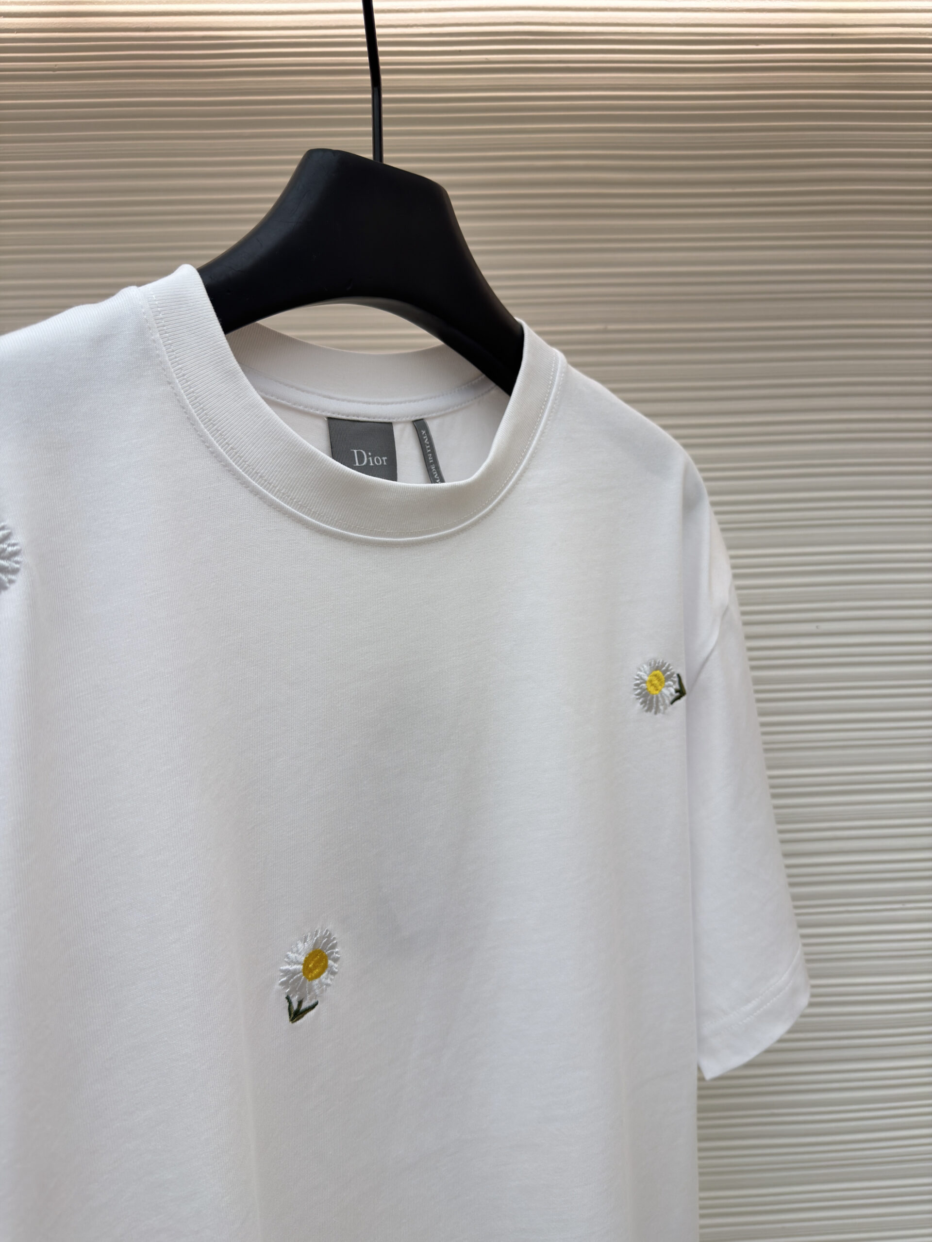 A premium Dior White Daisy Embroidered T-Shirt hanging on a black hanger, showing the front view with scattered daisy motifs and a matching back view.