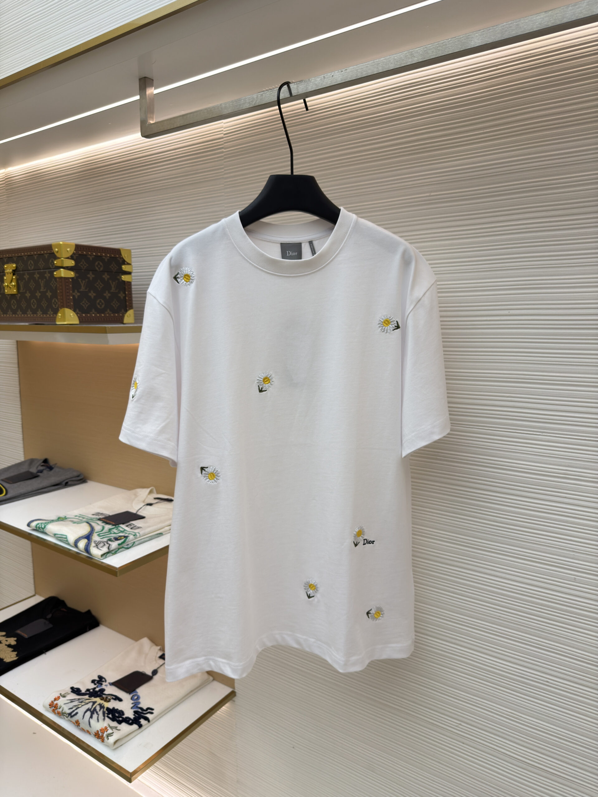 A premium Dior White Daisy Embroidered T-Shirt hanging on a black hanger, showing the front view with scattered daisy motifs and a matching back view.