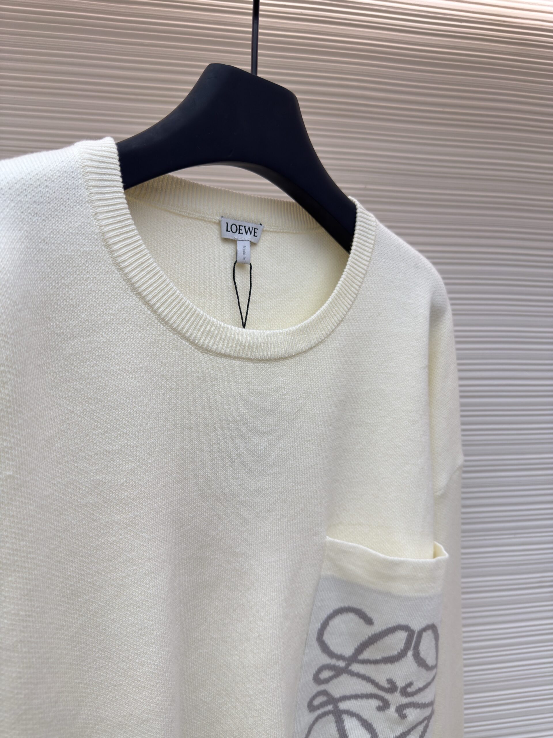 Front view of an oversized off-white cream Loewe knit sweater featuring a grey Anagram logo pocket hanging on a boutique display rack.
