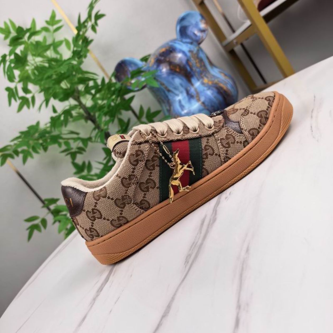 A side view of a Gucci low-top sneaker in beige GG Supreme canvas with the signature green and red Web stripe on a marble surface.