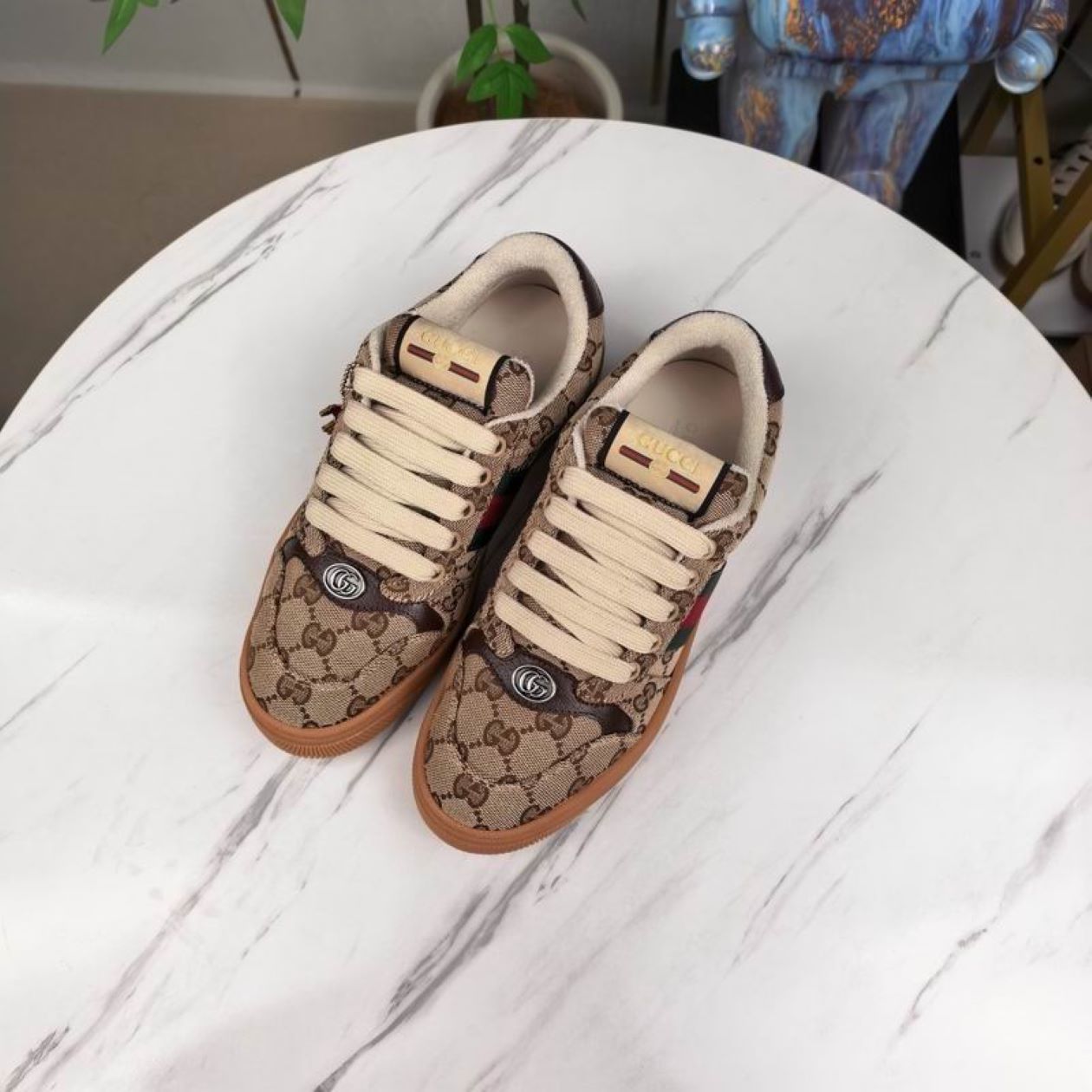 A side view of a Gucci low-top sneaker in beige GG Supreme canvas with the signature green and red Web stripe on a marble surface.