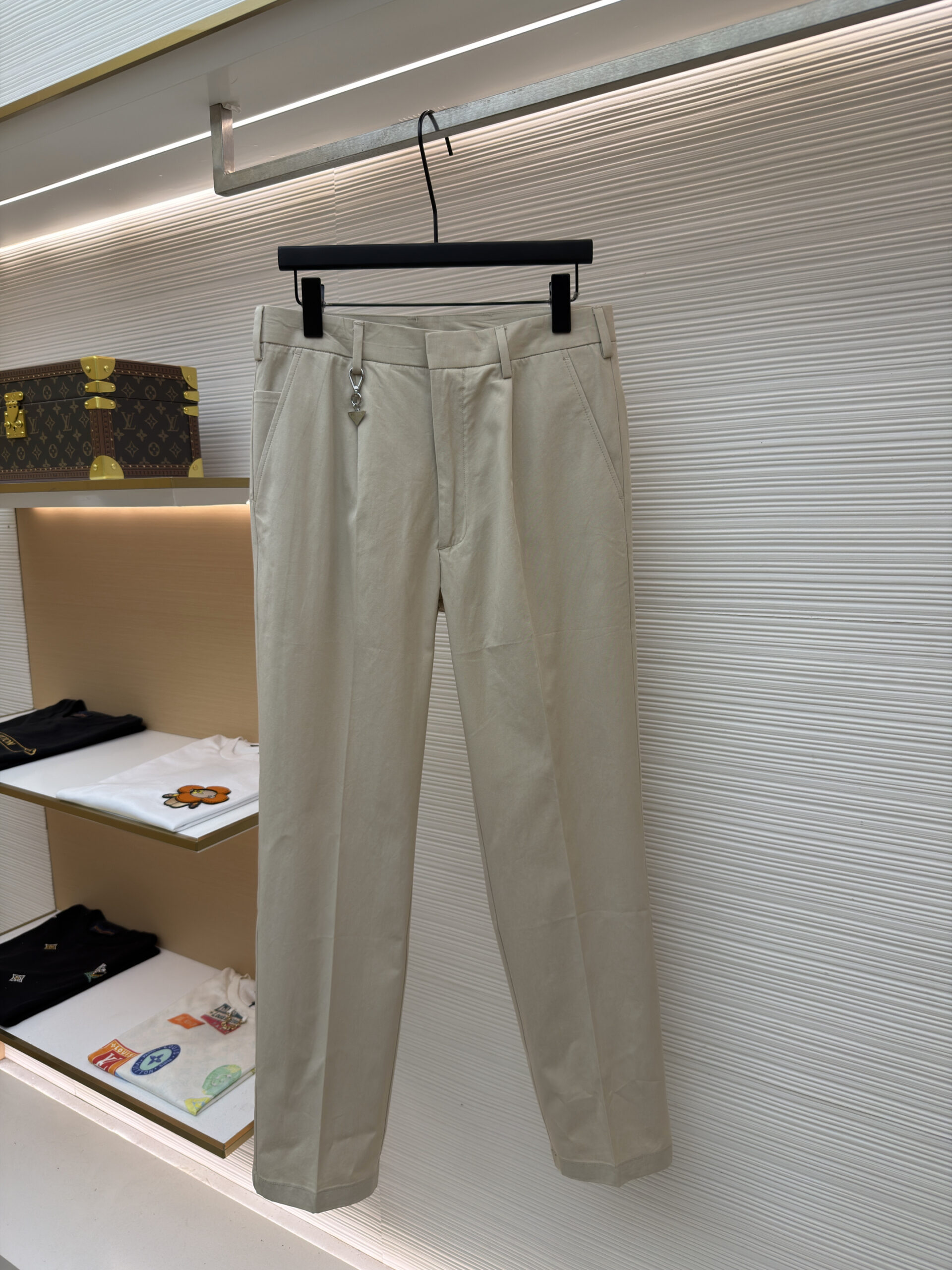 Front and back view of the premium Prada Logo Charm Beige Chino Trousers hanging on display, showcasing the tailored leg crease and rear buttoned pockets.