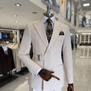 Mannequin displaying a modern beige men's suit with a side-buckle belt closure, peak lapels, dark paisley tie, and matching pocket square.