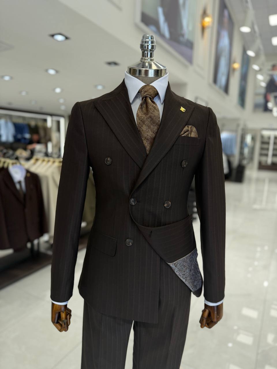 Mannequin showcasing a men's dark brown pinstripe double-breasted suit with an asymmetric wrap detail, a brown textured tie, and a white shirt.